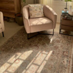 The Bustan Rug with intricate floral patterns in beige, blue, and red tones, styled in a cozy corner with a blush accent chair and wooden furniture.