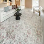 The Bustan Rug with elegant floral motifs in beige, blue, and red tones, styled in a modern open-concept living room with a white sofa and minimalist decor.
