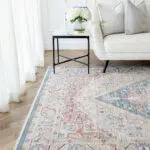 The Ishtar Medallion Rug in Dusty Azure displayed in a stylish living room setup with a white sofa and floral decor.