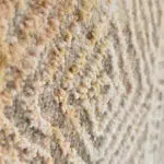 A detailed close-up of the Babylon Rug, showcasing its plush 480 reeds, 1600 density texture with soft beige, golden, and ivory fibers woven in an intricate geometric pattern.